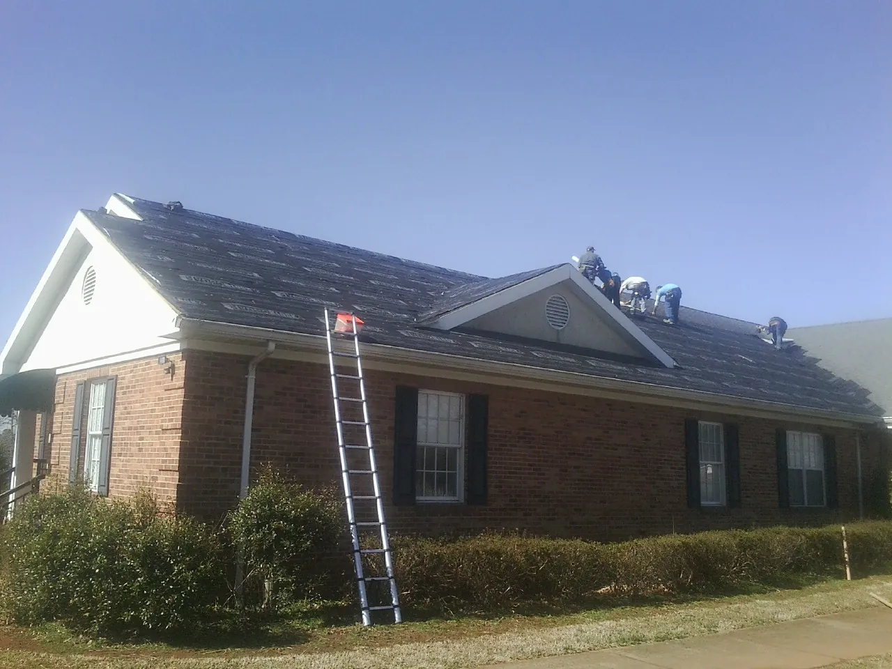 Crew tearing shingles off a brick ranch roof with a ladder set — Metal Roof Repair work by our crew in Drake